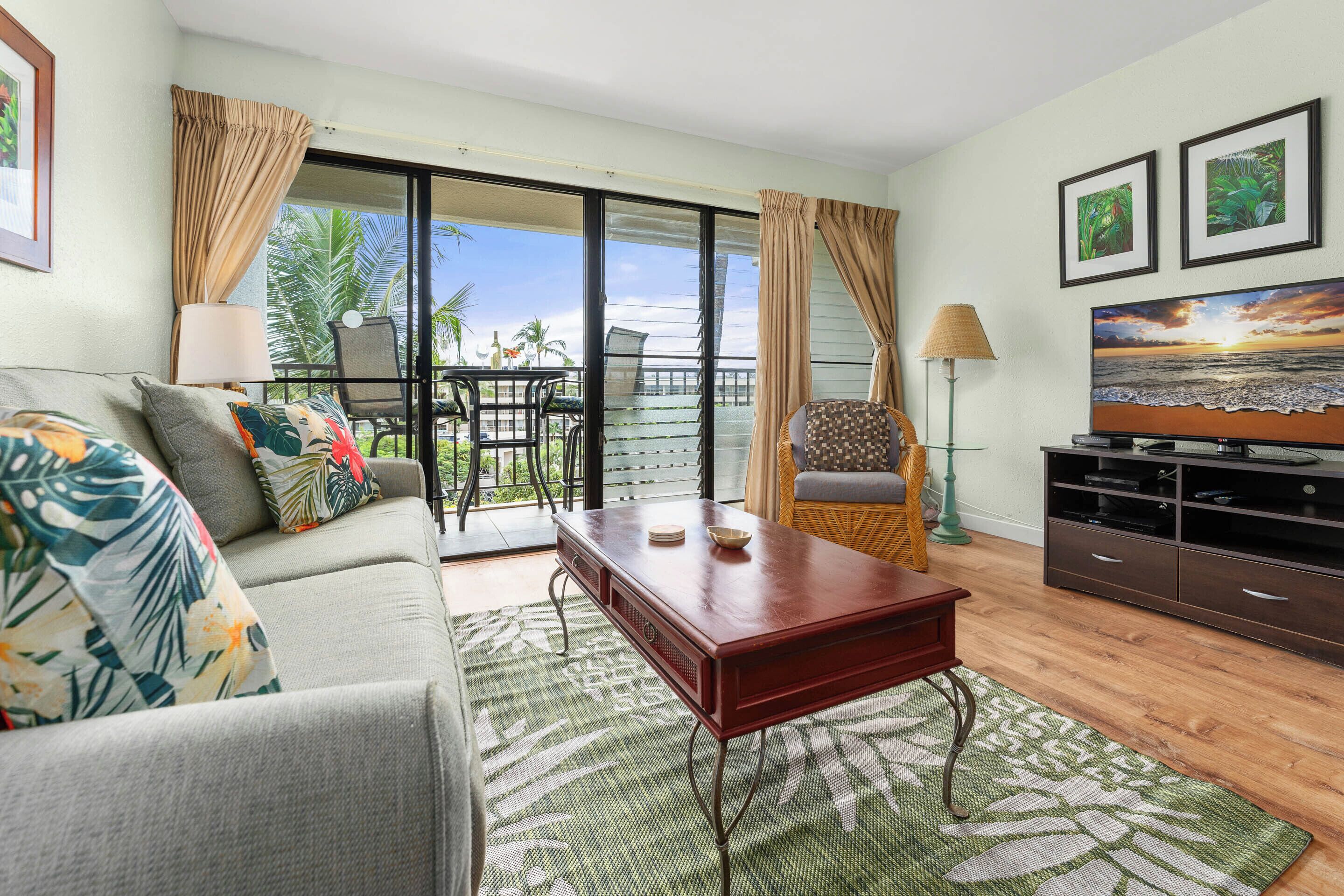 A living room with a sofa, coffee table, and TV stand, featuring a balcony with a view of palm trees and the ocean.