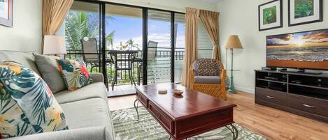 A living room with a sofa, coffee table, and TV stand, featuring a balcony with a view of palm trees and the ocean.