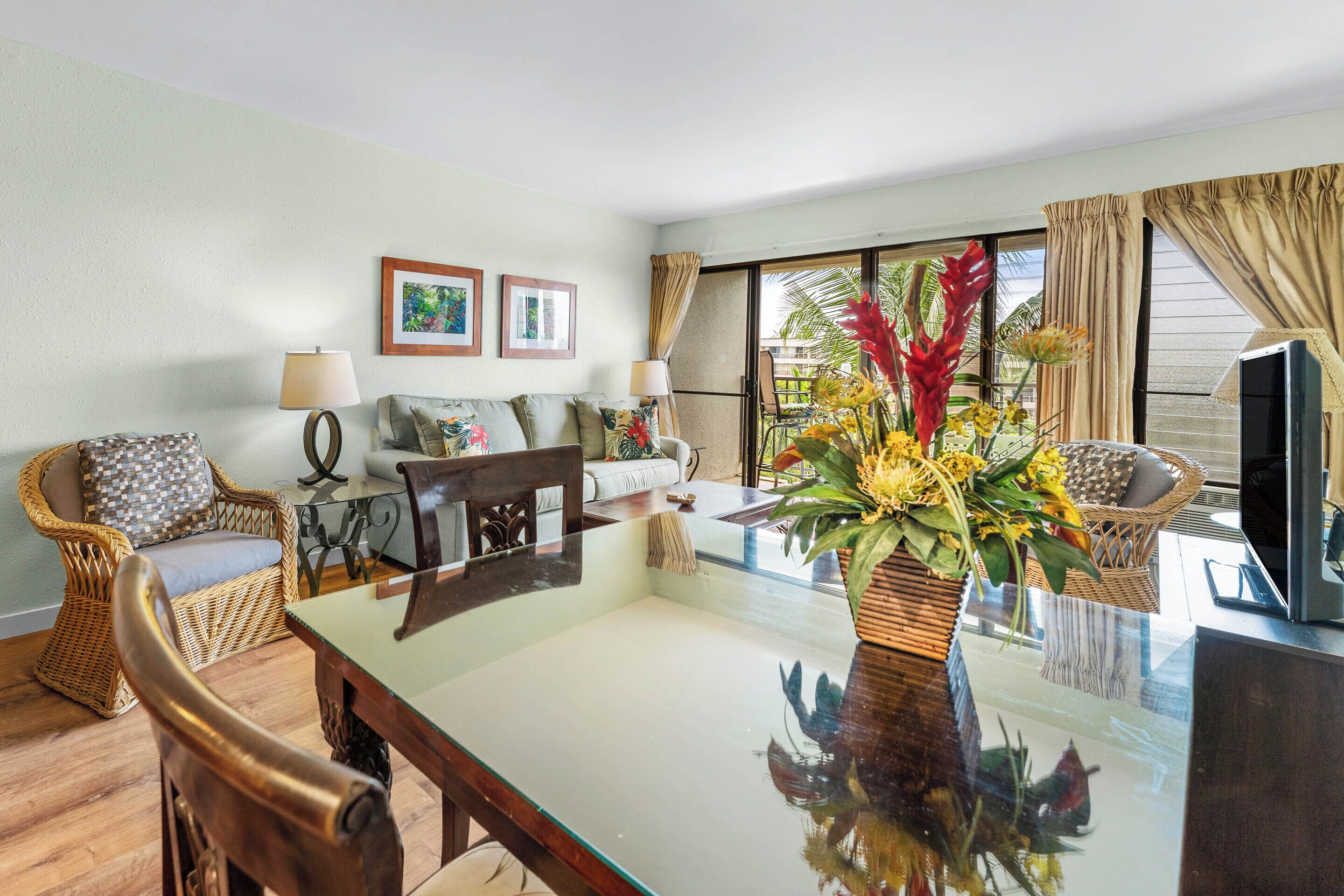 A living room with a glass-top table, wicker furniture, a TV, and a large window with curtains.
