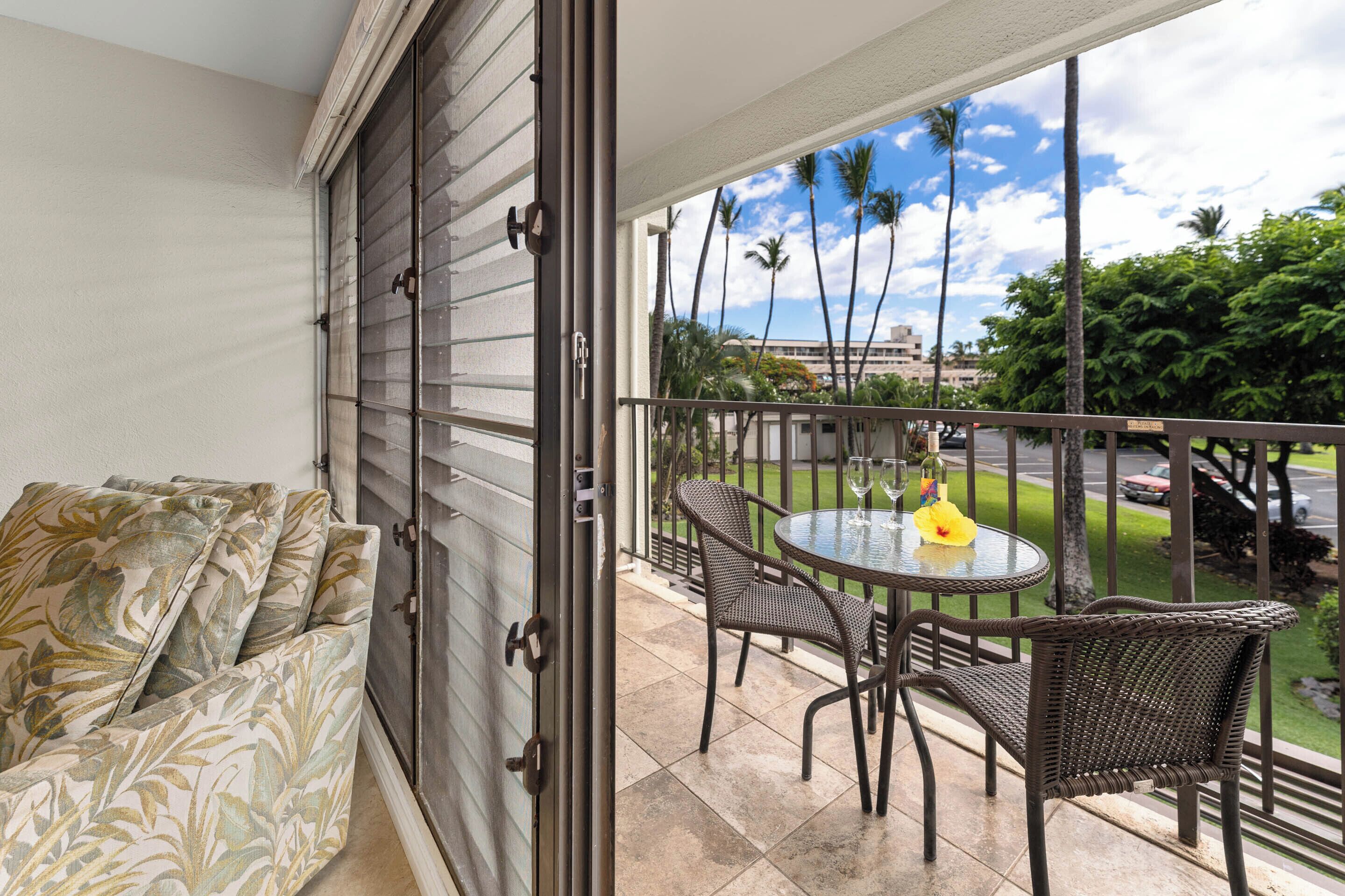 Un balcon avec vue sur des palmiers et une route. On y trouve une table ronde avec des chaises et un plateau en verre.