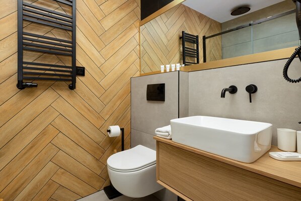 A modern bathroom with a wooden herringbone floor, a white toilet, a large mirror, a white sink, and a towel rack.