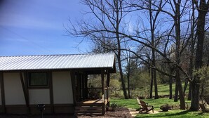 A single-story building with a metal roof, a porch with chairs, and a grassy yard with trees. - The Makenzee at Ridgeview. Asheville rental! Luxurious Glamping! (Alexander)