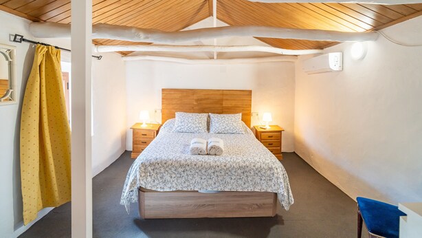Un lit bien fait, avec une couvre-lit à motifs floraux et deux serviettes enroulées, une tête de lit en bois, des lampes de chevet et un rideau jaune.