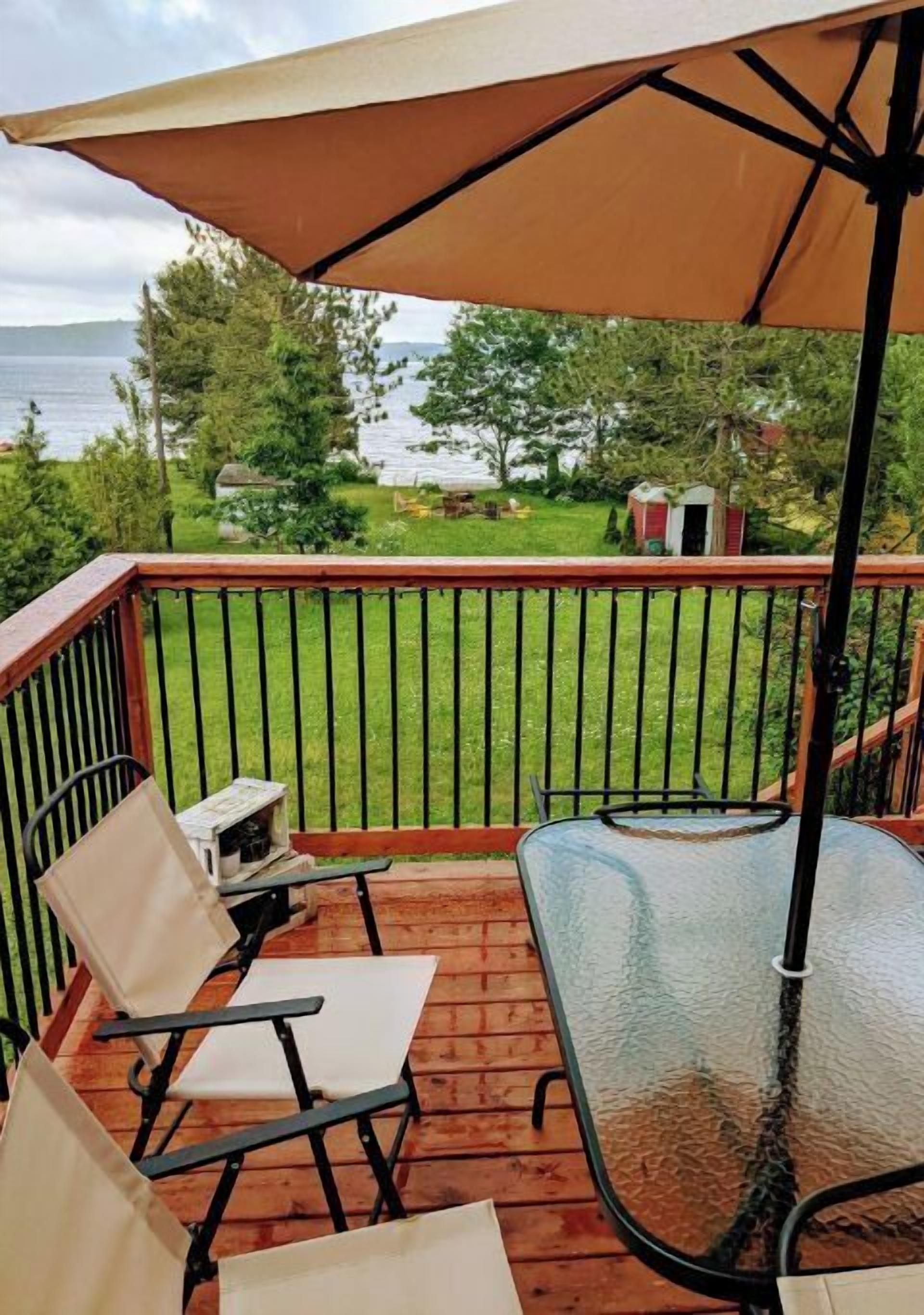 A deck with a glass table and chairs, a large umbrella, and a view of a lake and trees.