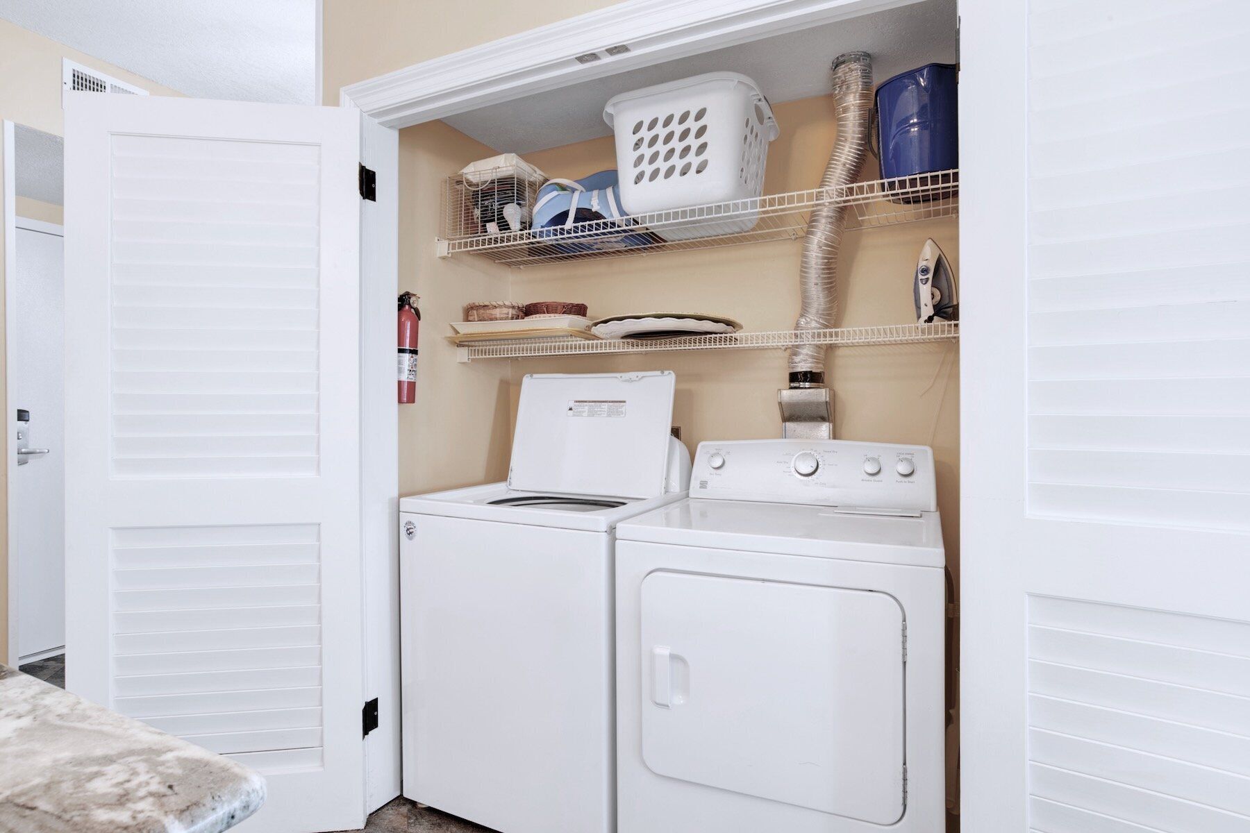 Une buanderie équipée de deux machines à laver et deux sèche-linge superposés, avec des étagères au-dessus pour ranger des paniers et des produits de nettoyage, et un extincteur fixé au mur.