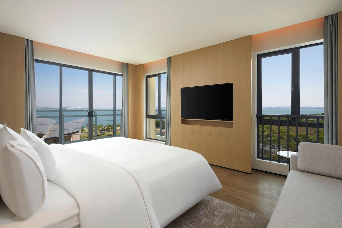 A hotel room with a large bed, a flat-screen TV, a balcony with a view of the city, and a sofa.