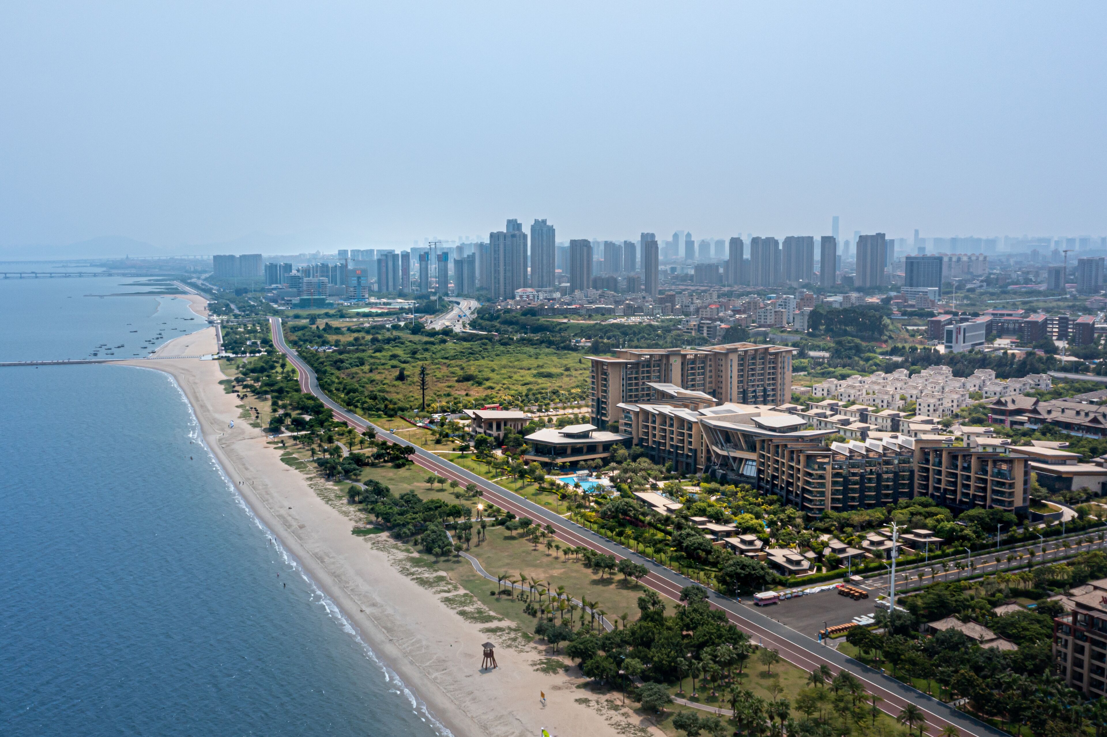 An aerial view of a coastal city with a beach, high-rise buildings, and a road running parallel to the shore.