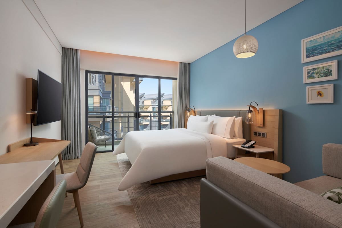 A modern hotel room with a large bed, a desk, a chair, a sofa, and a balcony with a view of buildings.