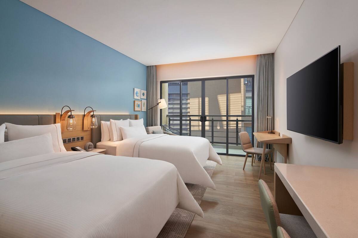 A hotel room with two beds, a desk, a chair, a TV, and a large window with a view of buildings.