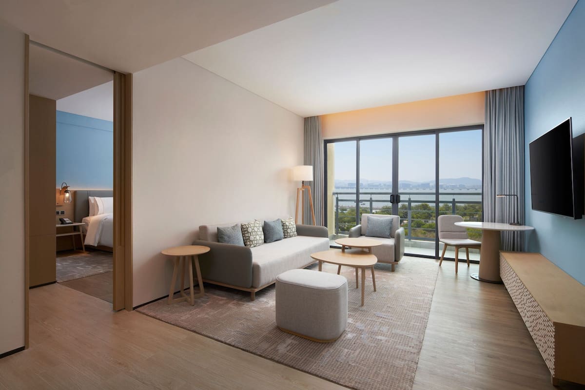 A modern hotel room with a large window, a sofa, a coffee table, a TV, and a small desk.