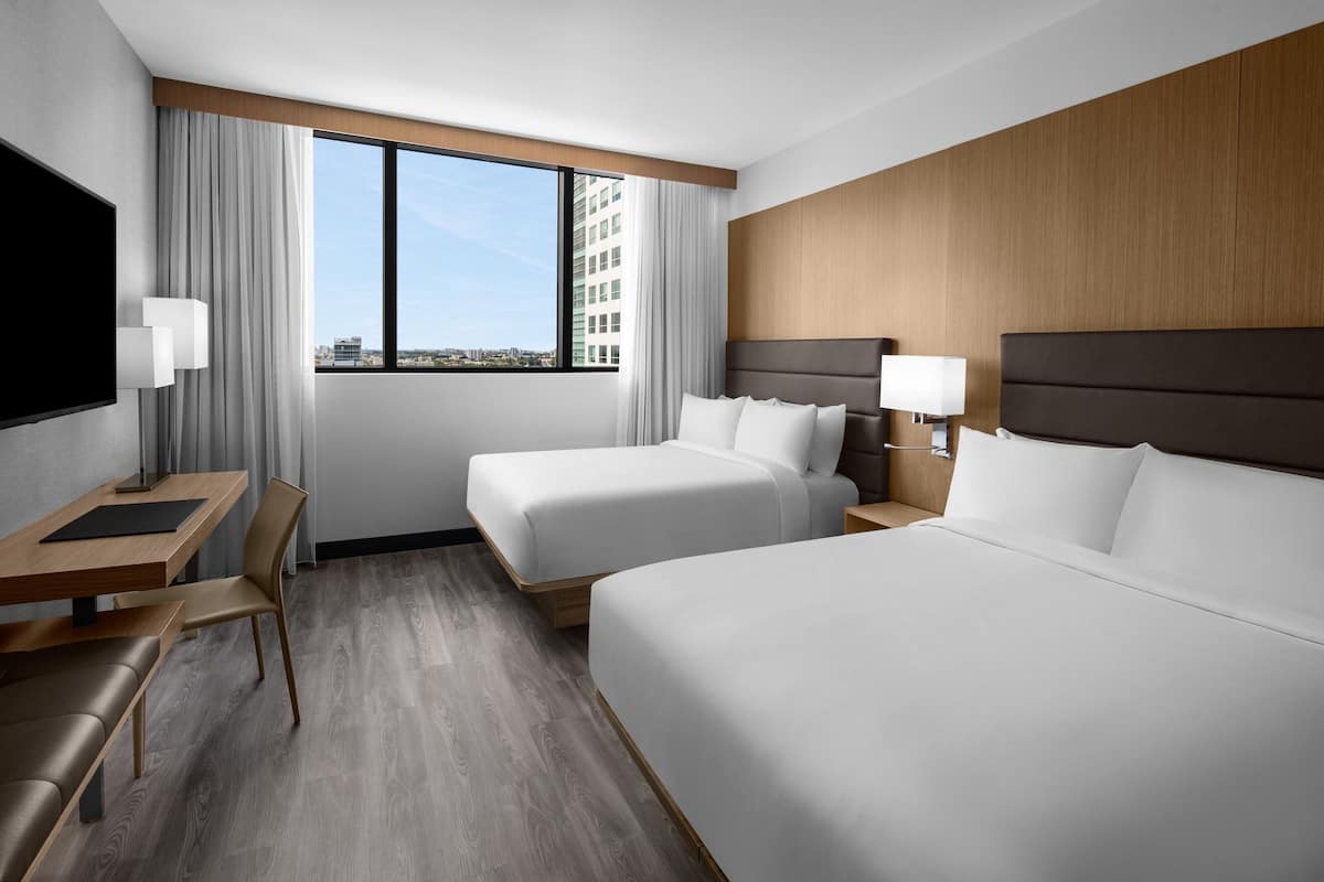 Ein Hotelzimmer mit zwei Betten, einem Schreibtisch, einem Stuhl, einem Fernseher und einem großen Fenster mit Blick auf die Stadt.