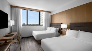A hotel room with two beds, a desk, a chair, a TV, and a large window with a city view.