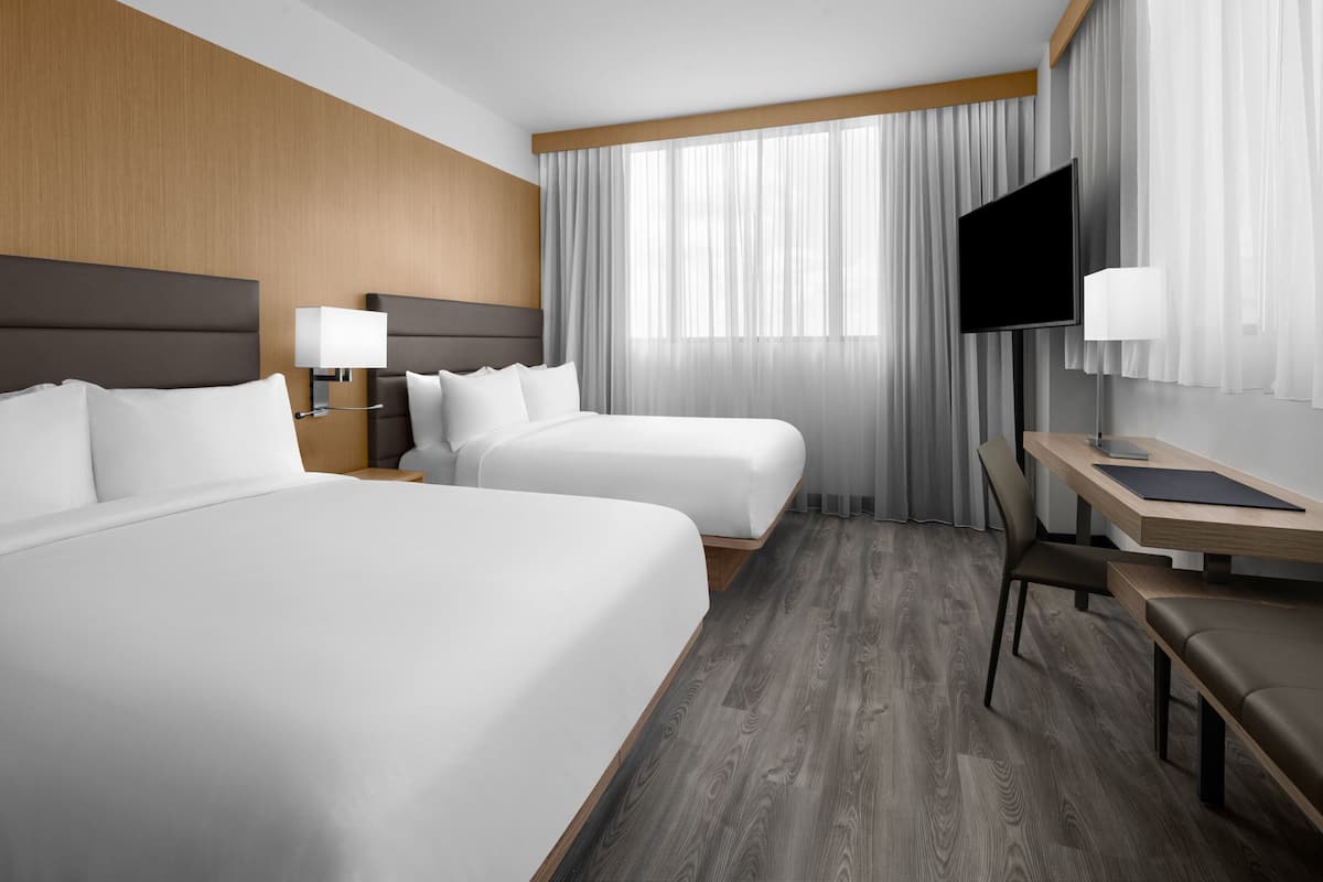 Ein Hotelzimmer mit zwei Betten, einem Schreibtisch, einem Sessel, einem Fernseher und einem Fenster mit Vorhängen.
