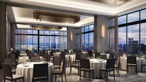 A spacious dining area with round tables and chairs, large windows offering a city view, and modern lighting fixtures.