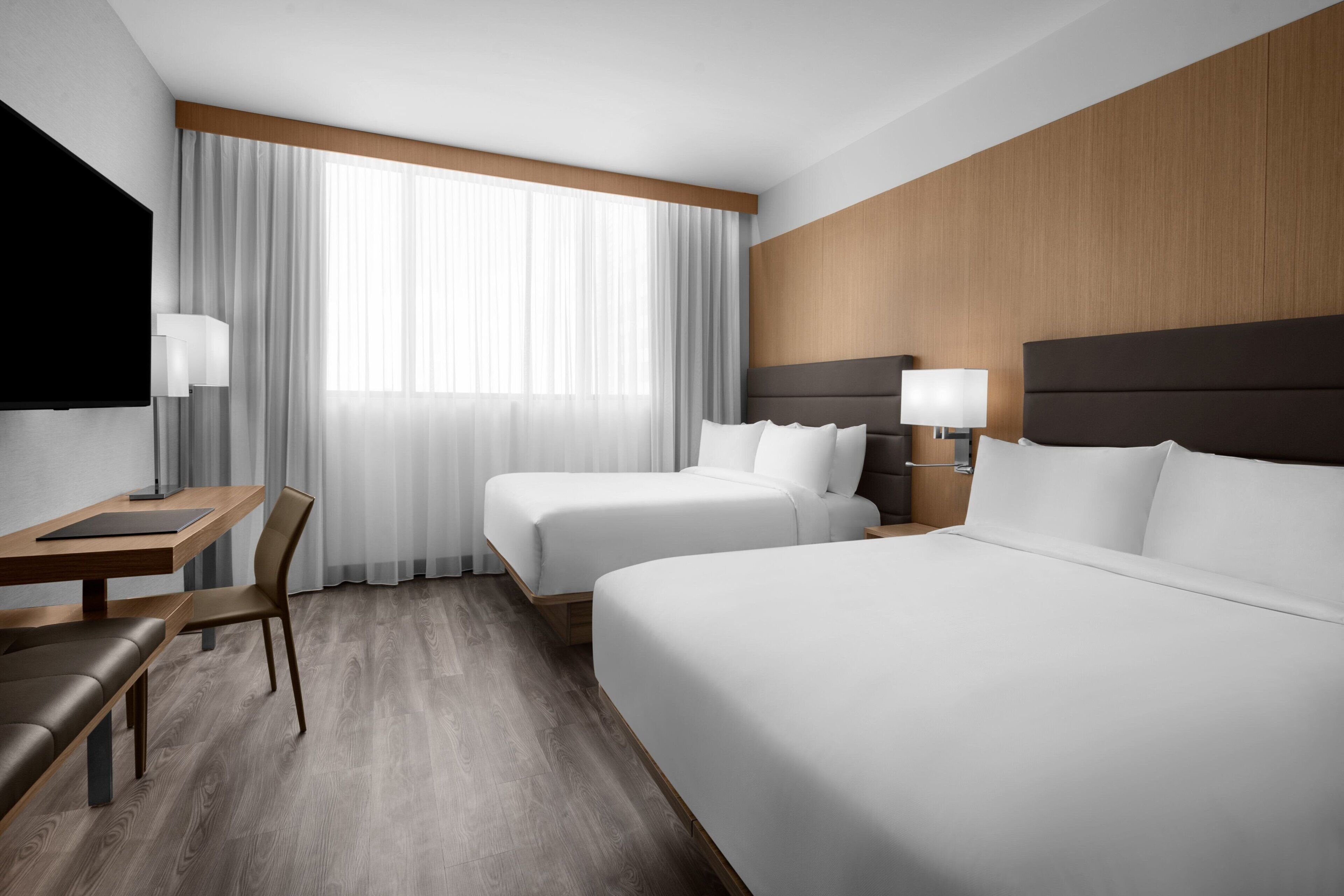 A hotel room with a large bed, a smaller bed, a desk with a chair, a TV mounted on the wall, and a window with curtains.
