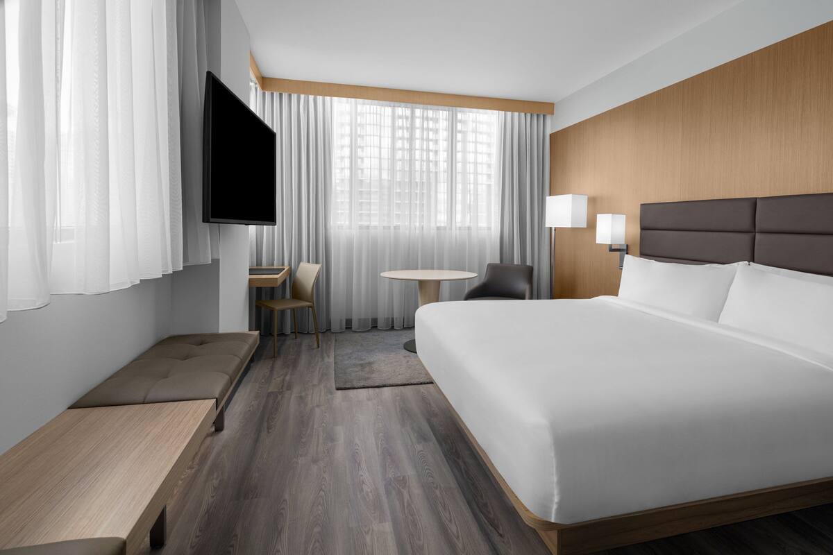 Ein modernes Hotelzimmer mit einem großen Bett, einem flachbildfernseher an der Wand, einem kleinen runden Tisch, einem Sessel und einem Schreibtisch.