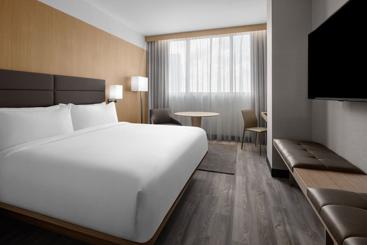 Ein modernes Hotelzimmer mit einem großen Bett, einer Sitzecke mit Tisch und Stühlen, einem Fernseher und einem Fenster mit Vorhängen.