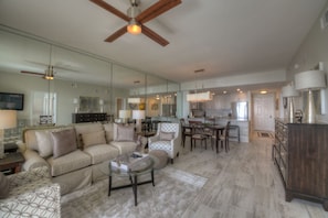 A spacious living room with a large sofa, a coffee table, and a dining area with chairs and a table. There is a kitchen in the background with cabinets and a refrigerator.