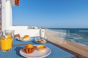 Tavola imbandita per la colazione con croissant, un muffin e un bicchiere di succo d'arancia su un balcone con vista sulla spiaggia.