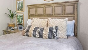 A neatly made bed with a wooden headboard, a bedside table with a 'WELCOME' sign, a potted plant, and framed botanical prints on the wall. - Downtown Caldwell | Golf Course View | Near I-84 | Fast WiFi | Sleeps 5 (Caldwell)