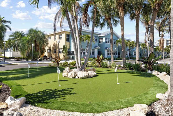 A well-maintained artificial green lawn with golf holes, surrounded by palm trees and rocks, with a building in the background.