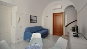 A living room with a blue sofa, a glass coffee table, a white armchair, a wooden door, and a small potted plant. - La Piazzetta by Amalfidreams (MINORI)