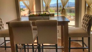 Un coin repas comprenant une table et quatre chaises, une fenêtre ornée de rideaux légers et une vue sur un paysage de montagnes et une piscine.