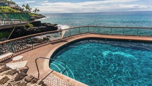 Ein Pool mit klarem, blauem Wasser, umgeben von einer Steindeck und einem Metallgeländer, mit Blick auf eine Küstenlinie mit Palmen und den Blick aufs Meer.
