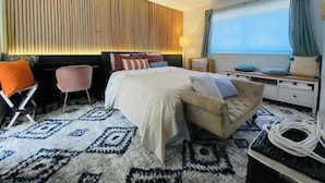 Ein modernes Hotelzimmer mit Bett, Nachttisch, Sessel und einem großen Fenster mit Vorhängen.