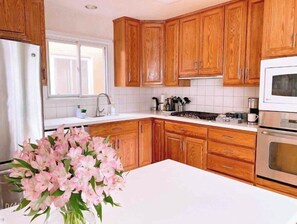 A kitchen with wooden cabinets, a white countertop, a stainless steel refrigerator, a microwave, and a vase of pink flowers.