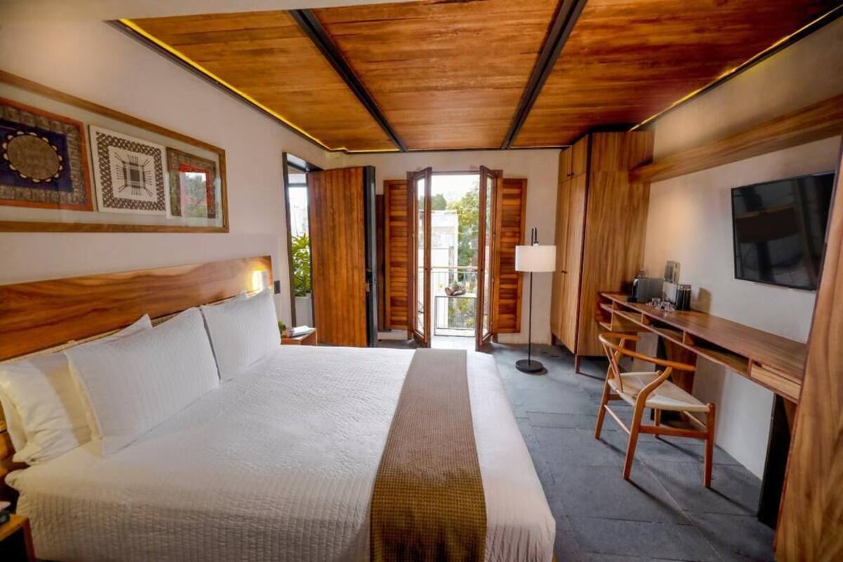 Habitación de hotel con una cama grande, cabecera de madera, un escritorio con silla, televisor y balcón con puertas abiertas.