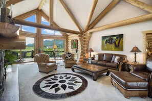 Ein geräumiges Wohnzimmer mit hohen Decken, Holzbalken, einem großen Fenster und Blick ins Freie.