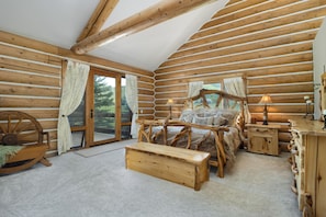 A log cabin bedroom with a wooden bed, bedside tables, a bench, and a view through a glass door.