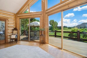 A log cabin interior with large windows, a wooden deck, and a view of a mountainous landscape.
