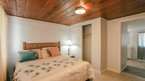 A bedroom with a wooden ceiling, a bed with floral bedding, a nightstand with a lamp, and a door leading to a bathroom.