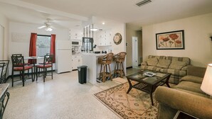 A spacious living area with a sofa, coffee table, and dining table with chairs. A kitchen area is visible in the background.