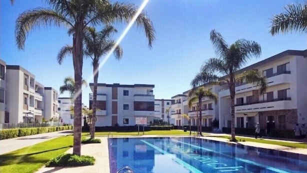 A swimming pool with a clear blue water surface, surrounded by modern buildings and palm trees. - Apartment Marbella beach 24/24 security (El Mansouria)