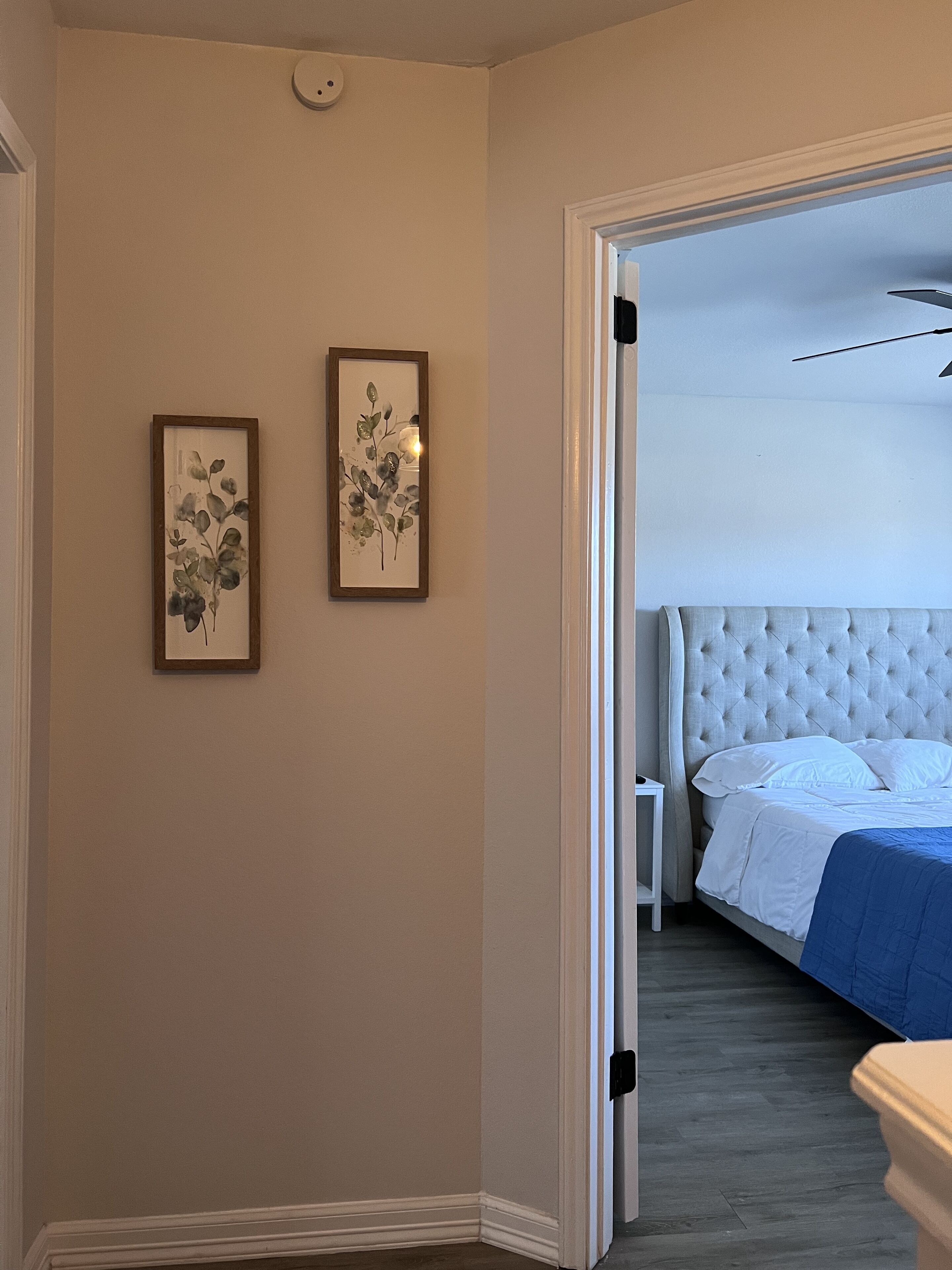 Une chambre avec une tête de lit capitonnée, un lit avec une literie blanche et une touche de couleur bleue. Deux œuvres d'art florales encadrées ornent le mur.