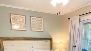 A bedroom with a large bed, a wooden bench, two wall-mounted light fixtures, and a view of the outdoors through a window with curtains. - Cozy and Secluded 2BR / 2BA fully equipped condo just a short walk to the beach  (Fernandina Beach)