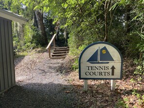A sign for Tennis Courts with an arrow pointing upwards, surrounded by trees and a gravel path.