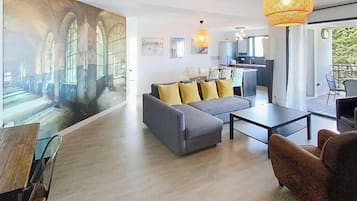 Un salon moderne avec un grand canapé, une table basse et un coin repas. On aperçoit une cuisine au fond, avec une grande fenêtre.