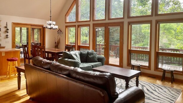A spacious living room with a high ceiling, large windows, a wooden dining table, and a leather sofa. - Spacious Cabin Right Next to Cacapon State Park (Berkeley Springs)