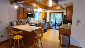A modern kitchen with wooden cabinets, a central island with bar stools, a stainless steel refrigerator, and a large window providing natural light. - Spacious Home in Waterville Estates close to the community center (Campton)
