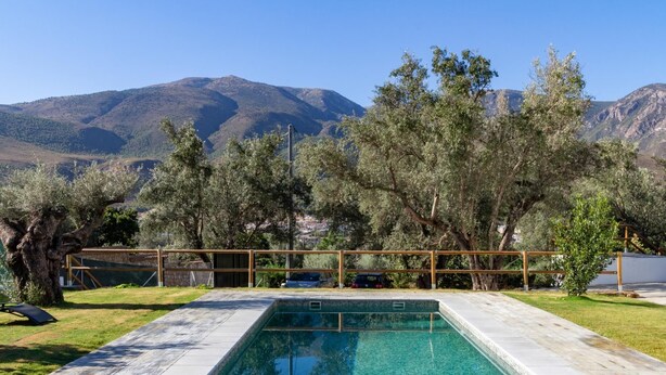 A rectangular swimming pool with clear blue water, surrounded by a wooden deck and a low fence. In the background, there are mountains and trees.