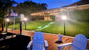 Eine Terrasse im Freien mit zwei Adirondack-Stühlen, einem kleinen Tisch und einem Mülleimer. Ein gepflegter Rasen und ein Gebäude mit Lichterketten sind im Hintergrund zu sehen.