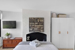 A bedroom with a white brick fireplace, a bed with white bedding, a wooden dresser, and a flat-screen TV on the wall. - Stylish Studio in Heart of Annandale Village (Annandale)