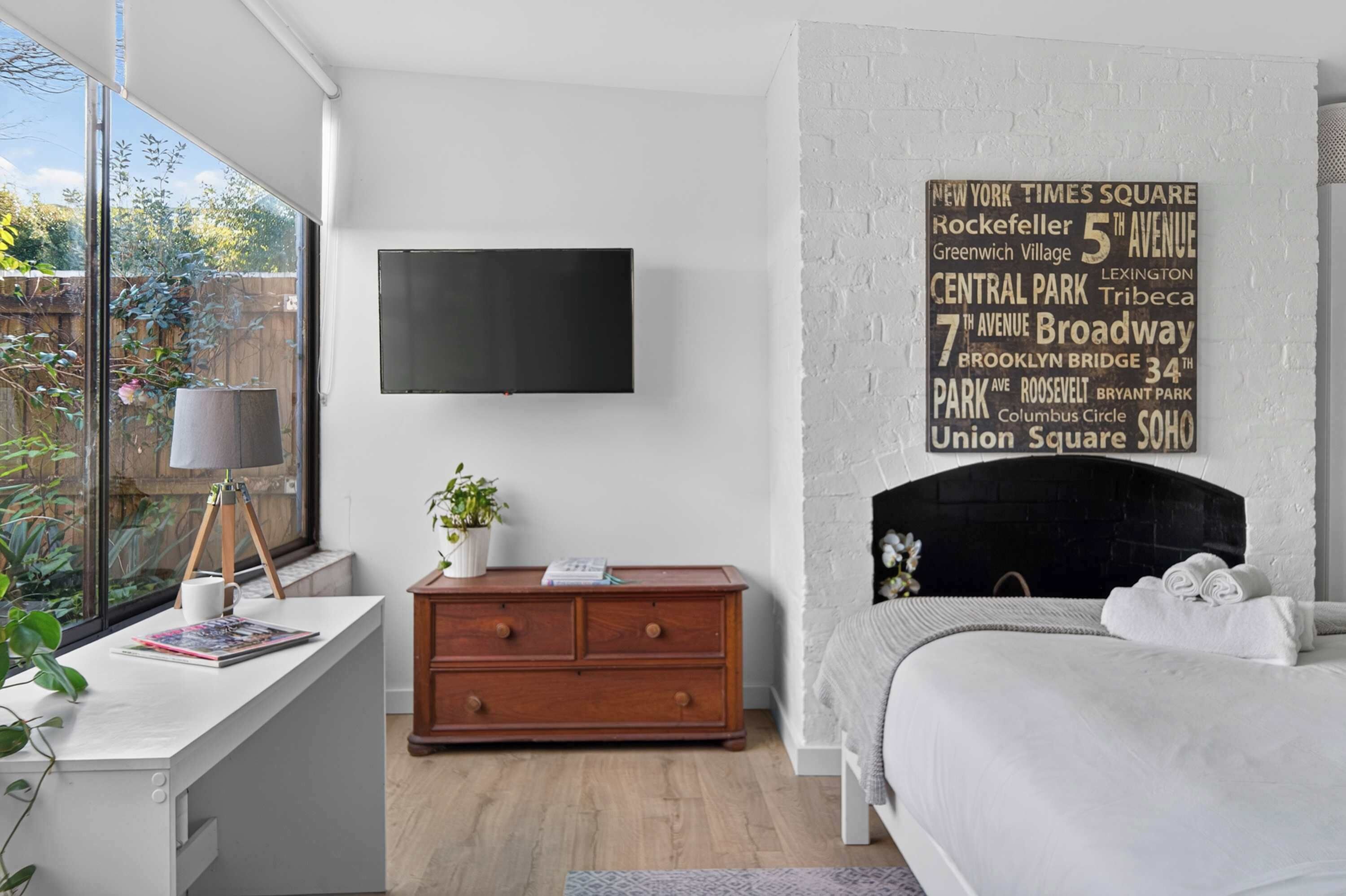 A bedroom with a white fireplace, a flat-screen TV mounted above it, a wooden dresser, and a window with a view of greenery.