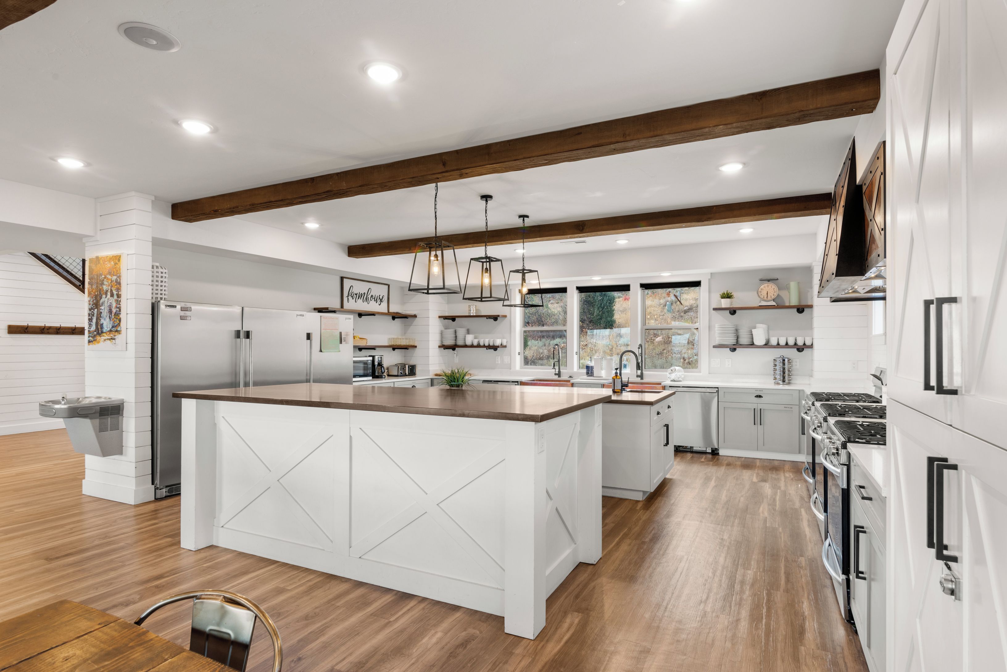 Une cuisine moderne dotée d’un grand îlot central, de meubles blancs, de poutres en bois et de luminaires suspendus.