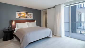 A modern hotel room with a large bed, a bedside table, a framed abstract artwork, and a view of a cityscape through a balcony.