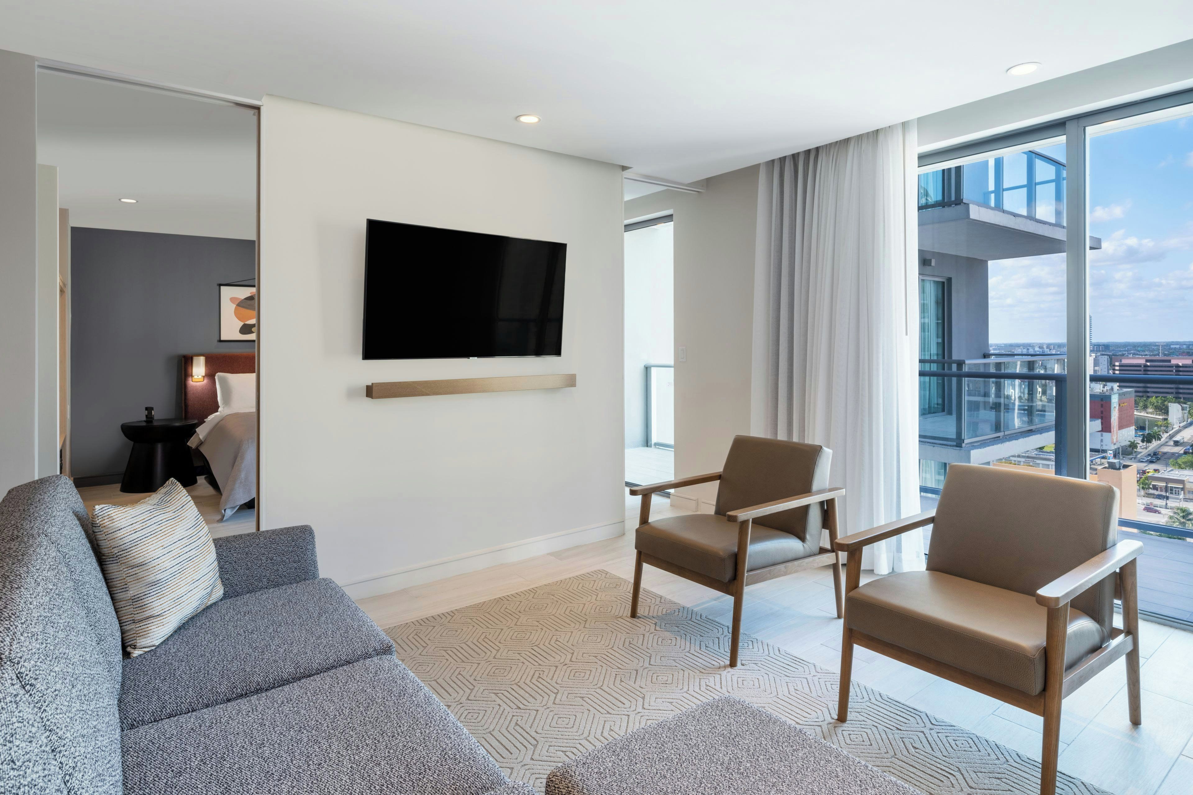 A modern living room with a flat-screen TV, two armchairs, a sofa, and a large window offering a city view.